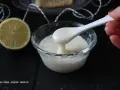 Una mano preleva con un cucchiaio una densa glassa al limone trasparente da una ciotola di vetro. Accanto, una fetta di limone.