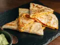 Deliziose quesadillas di pollo dorate e ripiene di formaggio filante, servite su una pietra scura con una ciotola di guacamole.