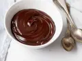 Una ciotola bianca piena di glassa al cioccolato e burro, lucida e invitante, con due cucchiai d'argento accanto.