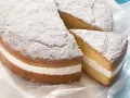 Una soffice torta paradiso con crema bianca, spolverata di zucchero a velo. Una fetta è stata tagliata, rivelando la deliziosa farcitura.
