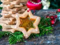 Biscotti a forma di stella con centro trasparente, decorati con piccoli fiocchi di neve, circondati da rami di abete e bacche rosse.