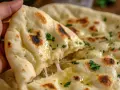 Una mano strappa un pezzo di naan al formaggio filante, cosparso di prezzemolo fresco e burro fuso.