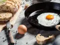 Un uovo sadzone sfrigola in padella, accanto a pane fresco e un uovo crudo.
