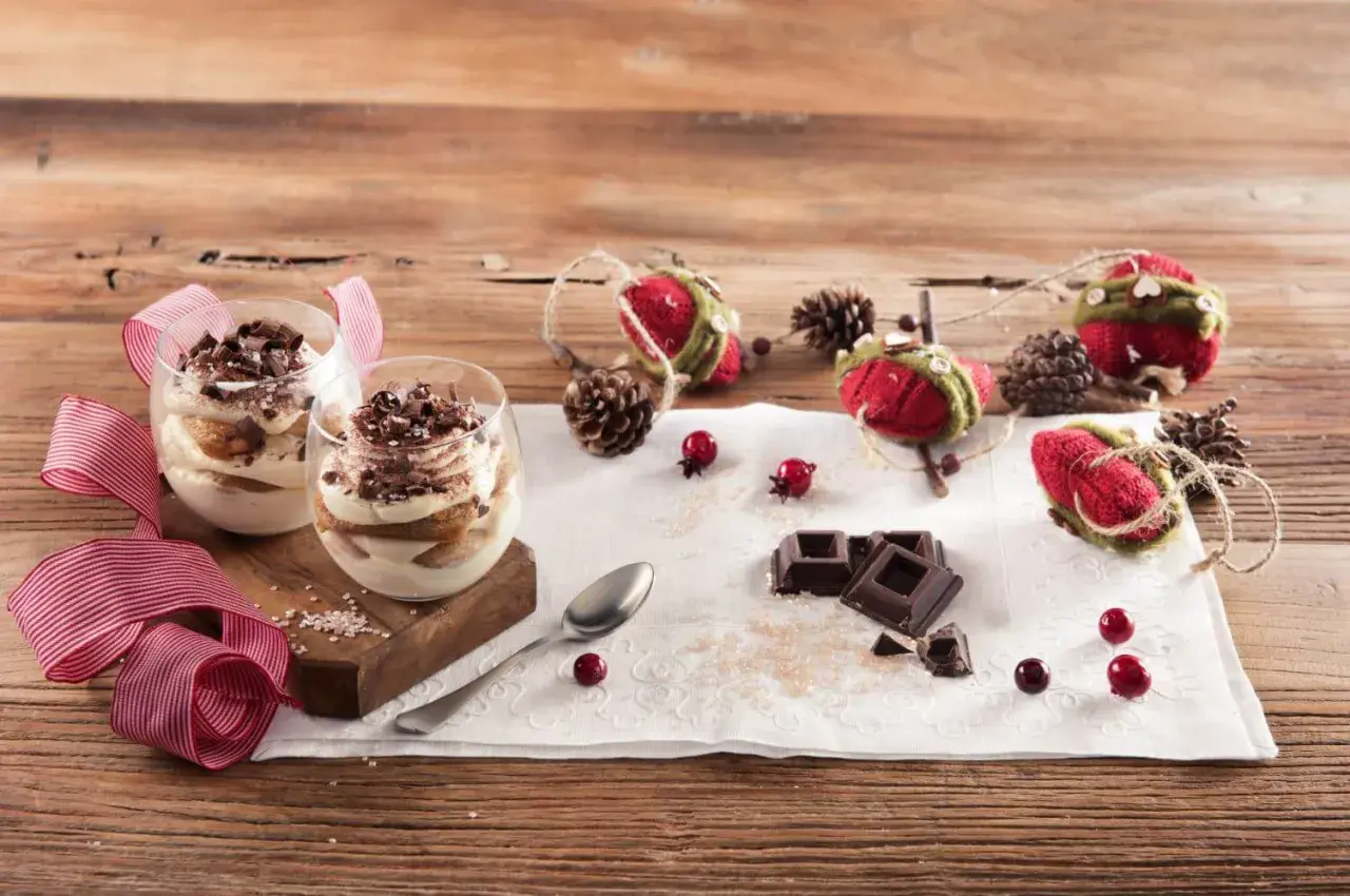 Due bicchieri di tiramisù Galbani, decorati con cioccolato e nastri rossi, su un tavolo di legno con decorazioni natalizie.