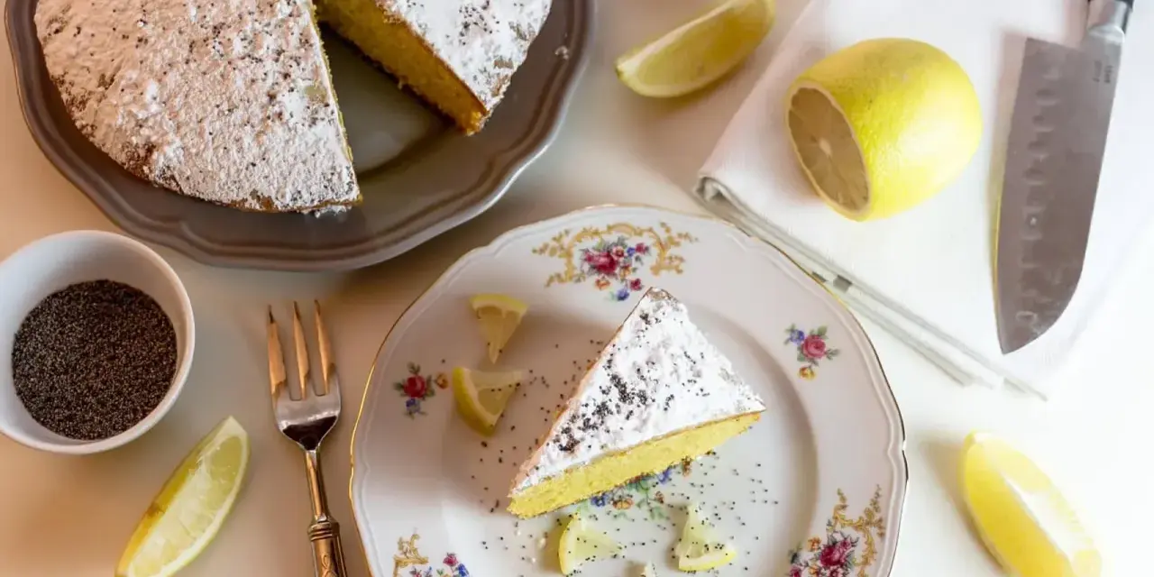 Una fetta di babka cytrynowa, cosparsa di zucchero a velo e semi di papavero, con spicchi di limone e una ciotolina di semi.