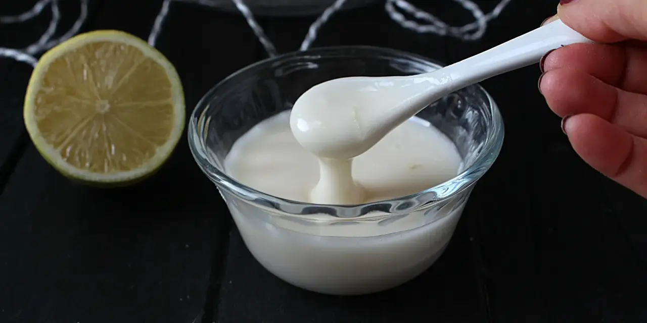 Una mano preleva con un cucchiaio una densa glassa al limone trasparente da una ciotola di vetro. Accanto, una fetta di limone.