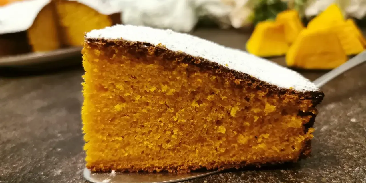 Una fetta di torta zucca senza glutine, soffice e dorata, cosparsa di zucchero a velo. Sullo sfondo, pezzi di zucca e un'altra torta.