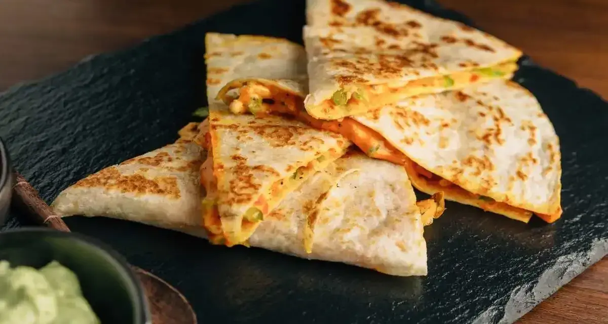 Deliziose quesadillas di pollo dorate e ripiene di formaggio filante, servite su una pietra scura con una ciotola di guacamole.