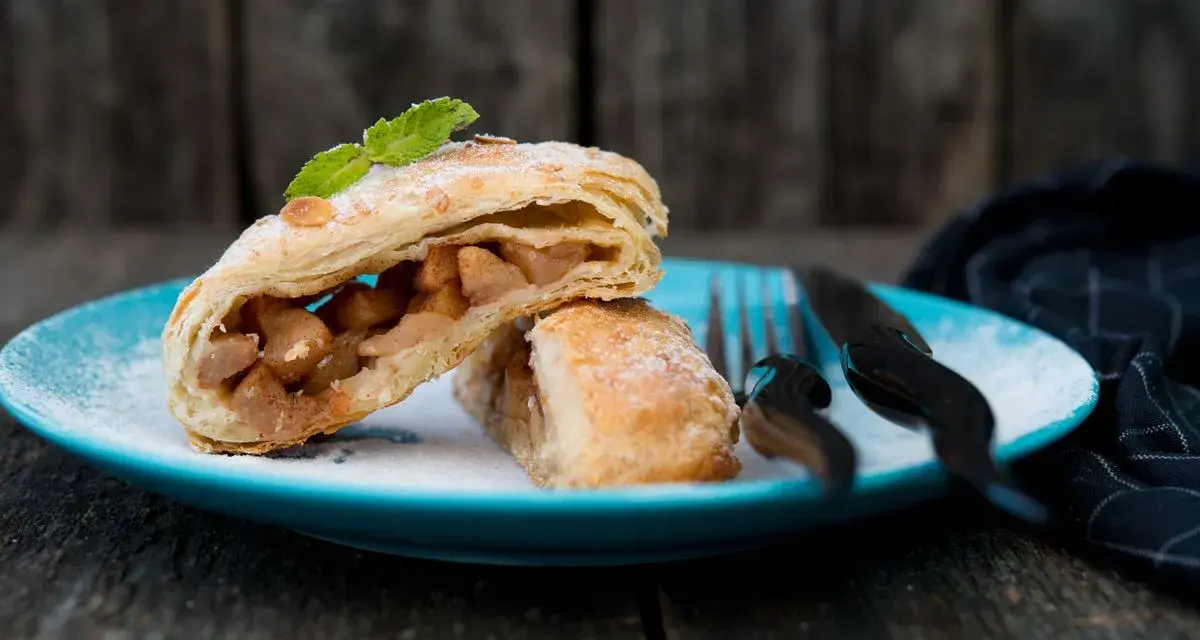Delizioso strudel pere cioccolato e amaretti, con ripieno di mele e un tocco di menta, su un piatto blu cosparso di zucchero a velo.