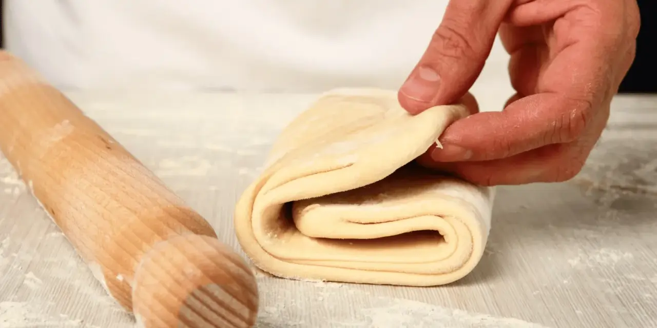 Mano che piega un impasto stratificato, pronto per diventare delizioso **ciasto francuskie**. Un mattarello è accanto.