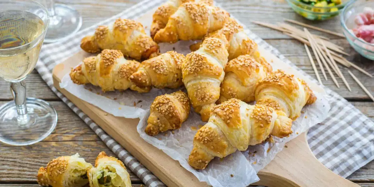 Croissant dorati con ripieno di pasta sfoglia e semi di sesamo, pronti per essere gustati con un bicchiere di vino.