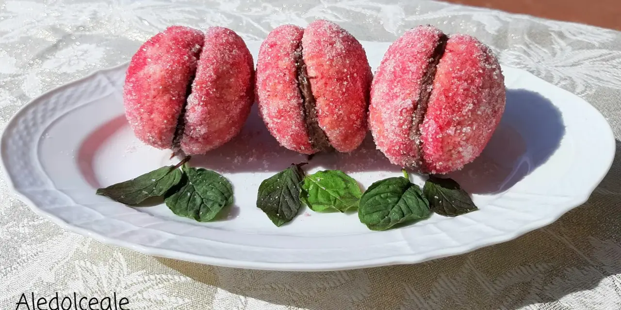 Tre dolci pesche abruzzesi ricoperte di zucchero, con un ripieno scuro, adagiate su un piatto bianco con foglie verdi.