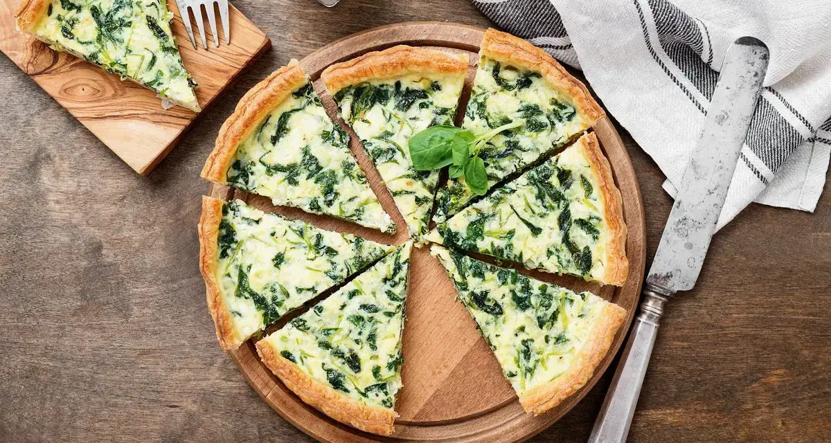 Torta salata spinaci e mozzarella, con un tocco di speck affumicato, tagliata a spicchi su un tagliere di legno.