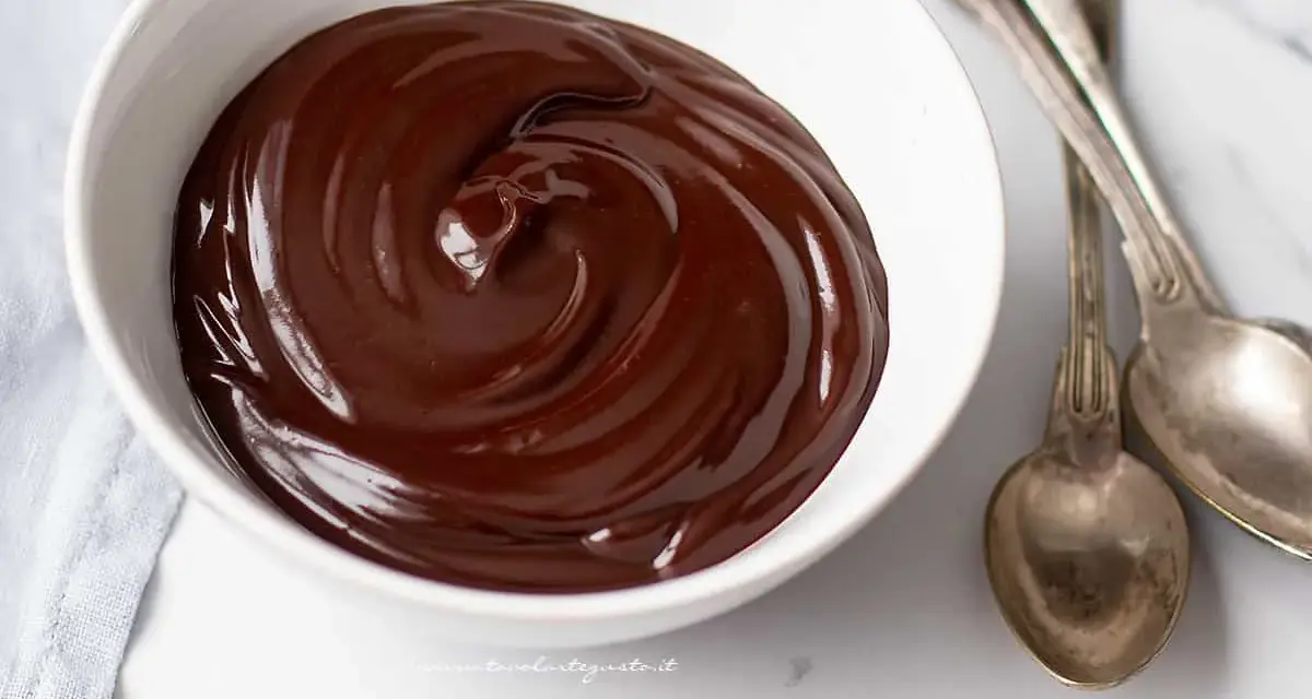 Una ciotola bianca piena di glassa al cioccolato e burro, lucida e invitante, con due cucchiai d'argento accanto.