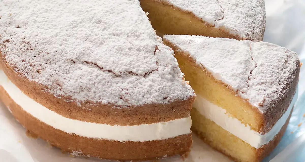 Una soffice torta paradiso con crema bianca, spolverata di zucchero a velo. Una fetta è stata tagliata, rivelando la deliziosa farcitura.