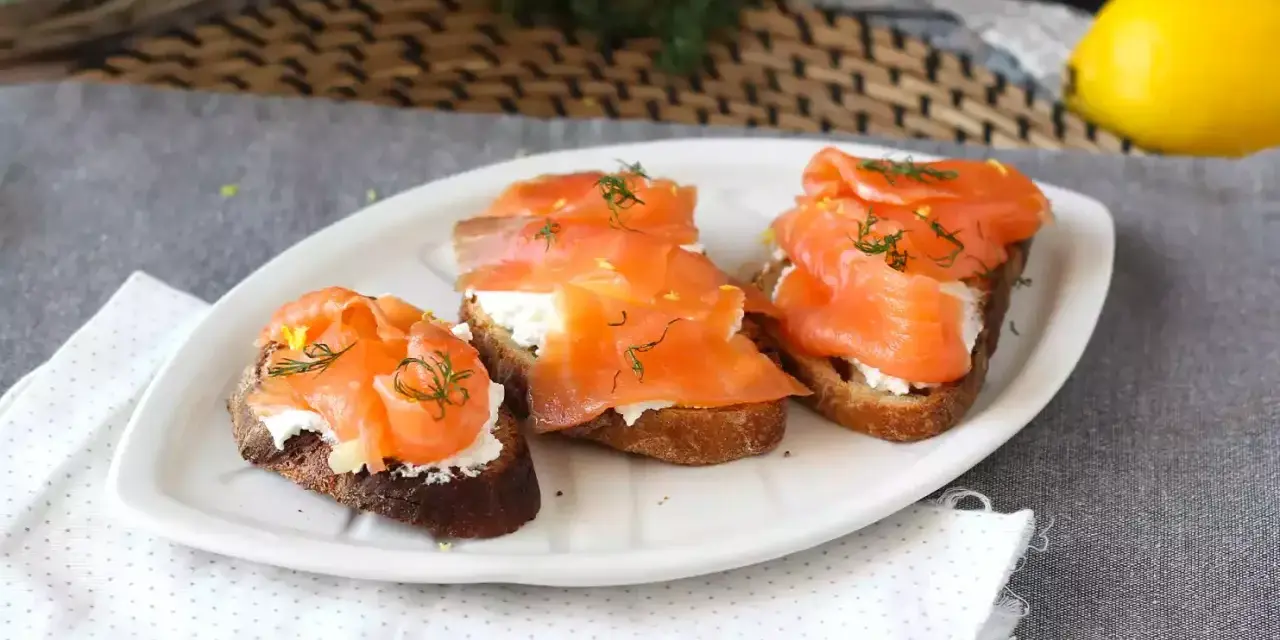 Tre bruschette con salmone affumicato, formaggio cremoso e aneto, guarnite con scorza di limone.