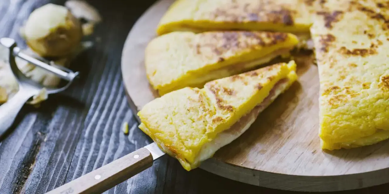 Focaccia di patate in padella con prosciutto cotto e formaggio, tagliata a spicchi su un tagliere di legno.