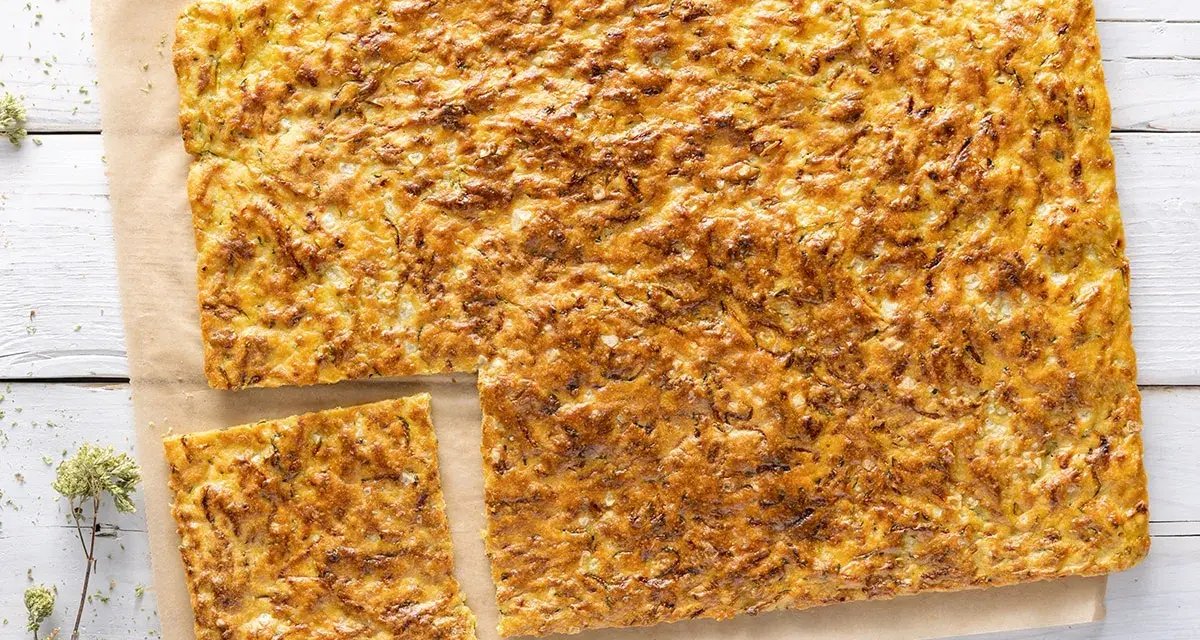 Schiacciata di zucchine e cipolle dorata e croccante, tagliata a pezzi su carta da forno.