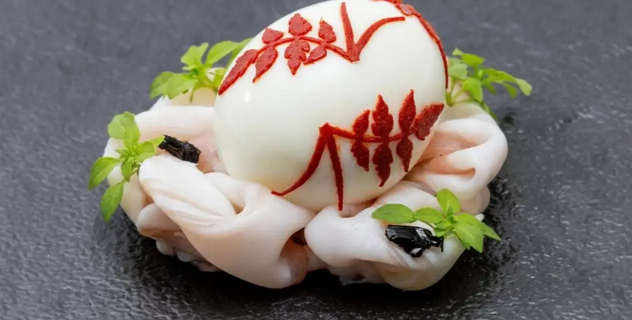 Uovo sodo decorato con motivi rossi, adagiato su un letto di calamari crudi e guarnito con erbe aromatiche. Un barzotto uovo elegante.