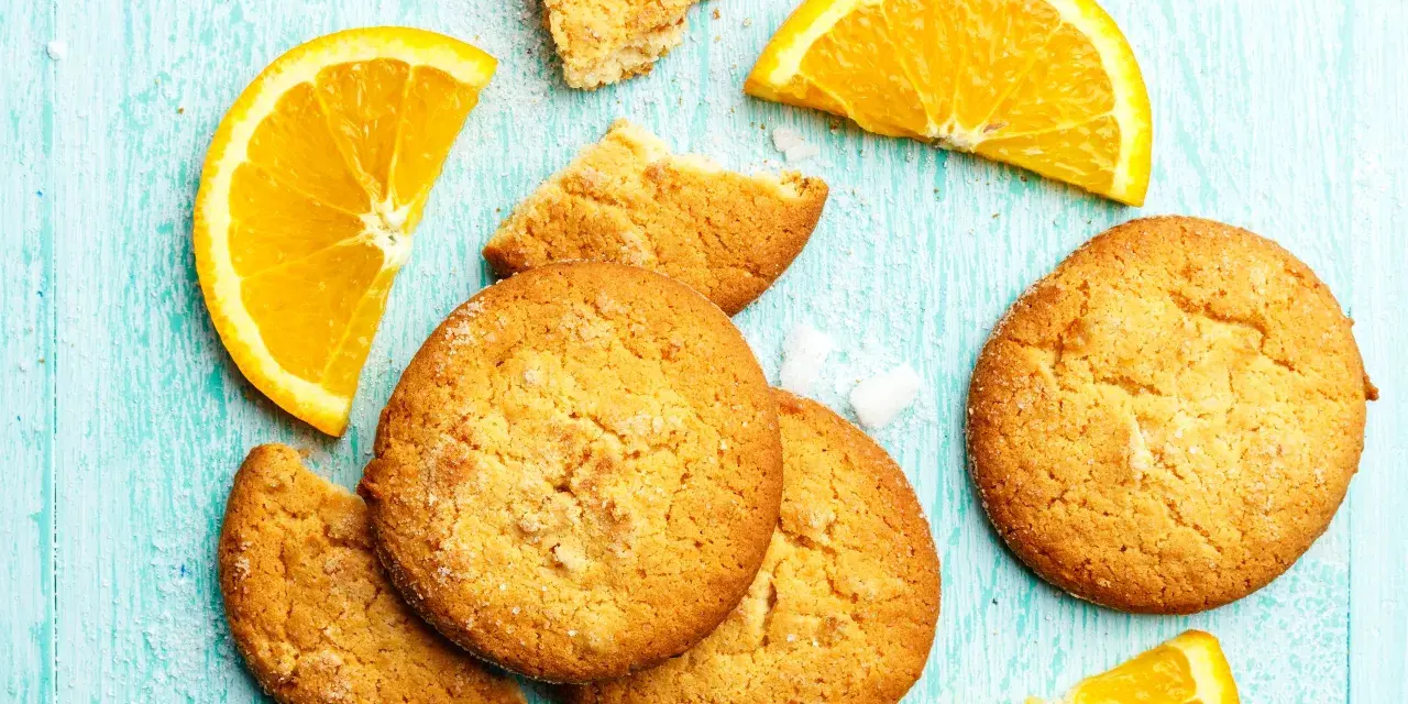 Biscotti all'arancia ricetta della nonna: dolci profumati e dorati, con spicchi d'arancia fresca su un tavolo di legno azzurro.