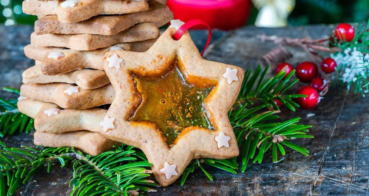Biscotti a forma di stella con centro trasparente, decorati con piccoli fiocchi di neve, circondati da rami di abete e bacche rosse.