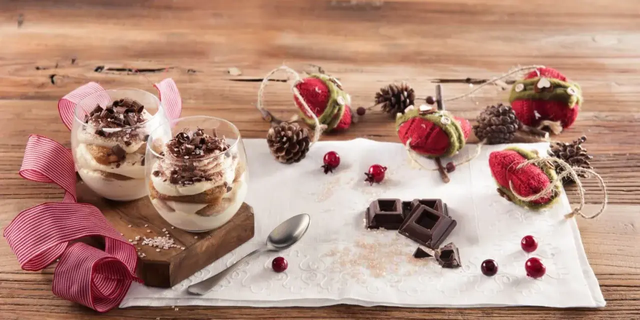 Due bicchieri di tiramisù Galbani, decorati con cioccolato e nastri rossi, su un tavolo di legno con decorazioni natalizie.