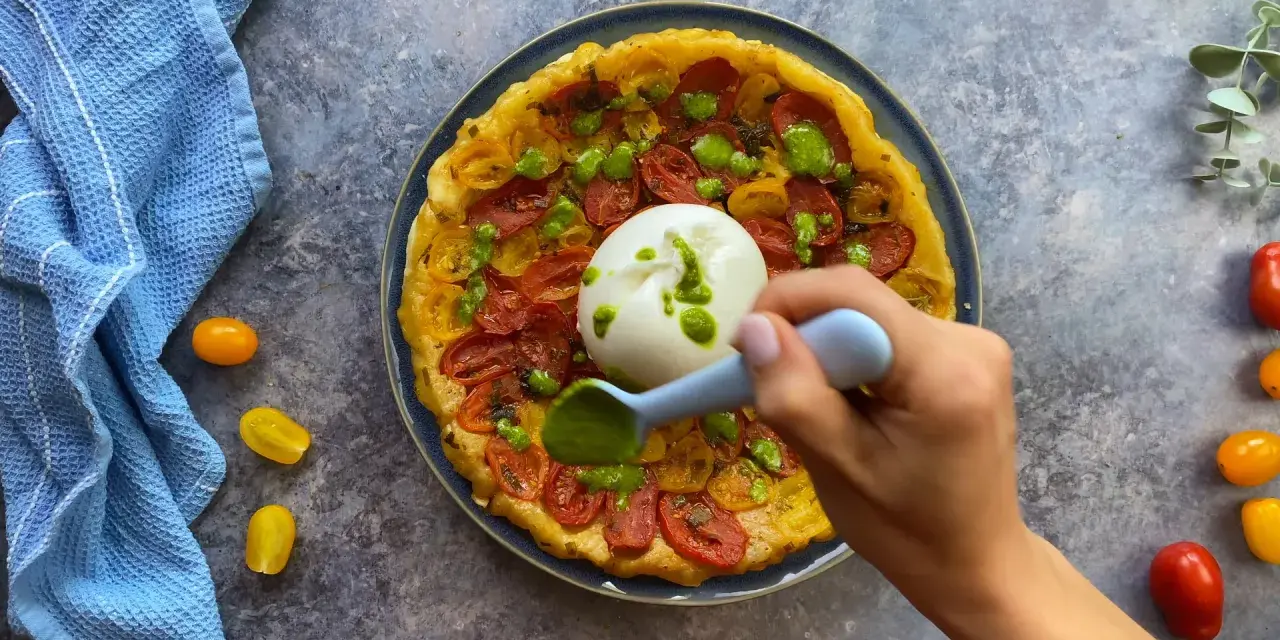 Tarte tatin pomodorini e burrata, con una mano che aggiunge un pesto verde.