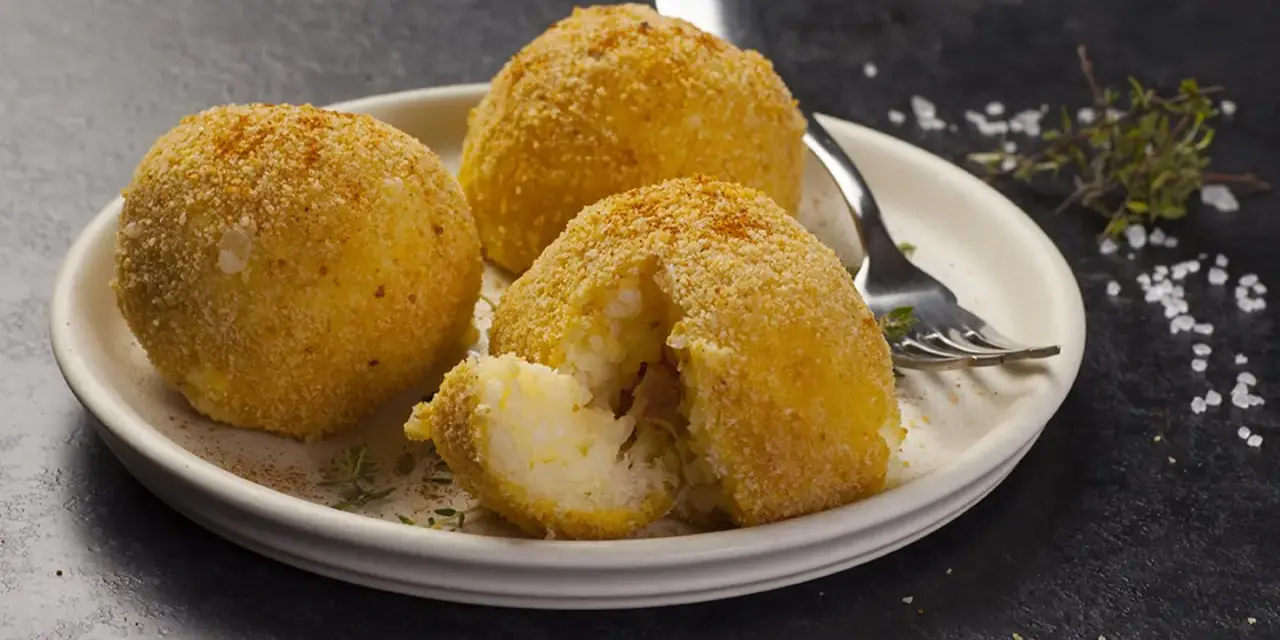 Tre arancini dorati su un piatto, uno aperto rivela il ripieno di riso. Un invito a provare il nostro arancini ricetta.