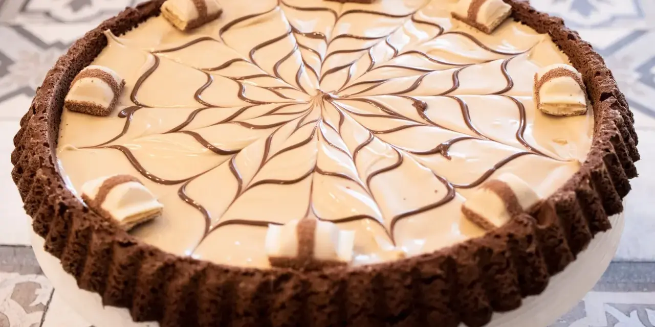 Torta con base croccante al cacao, ripiena di crema chiara e decorata con cioccolato fondente e pezzi di Kinder Bueno White.