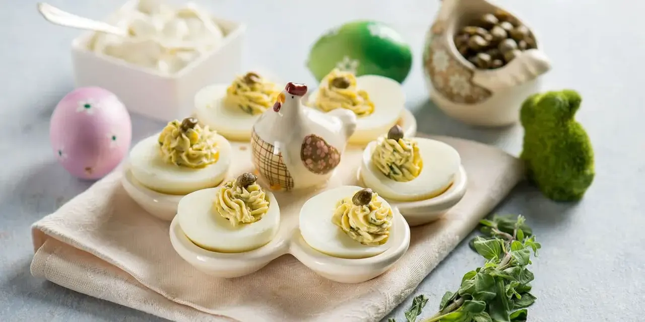 Uova ripiene vegetariane con ceci, guarnite con capperi e erbe aromatiche, su un piatto decorato con galline e uova pasquali.