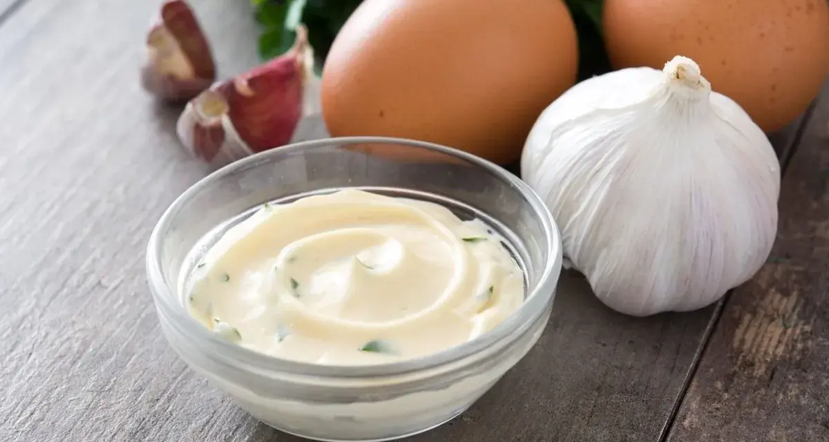 Una ciotola di cremoso sos aioli, con uova, aglio e prezzemolo sullo sfondo.