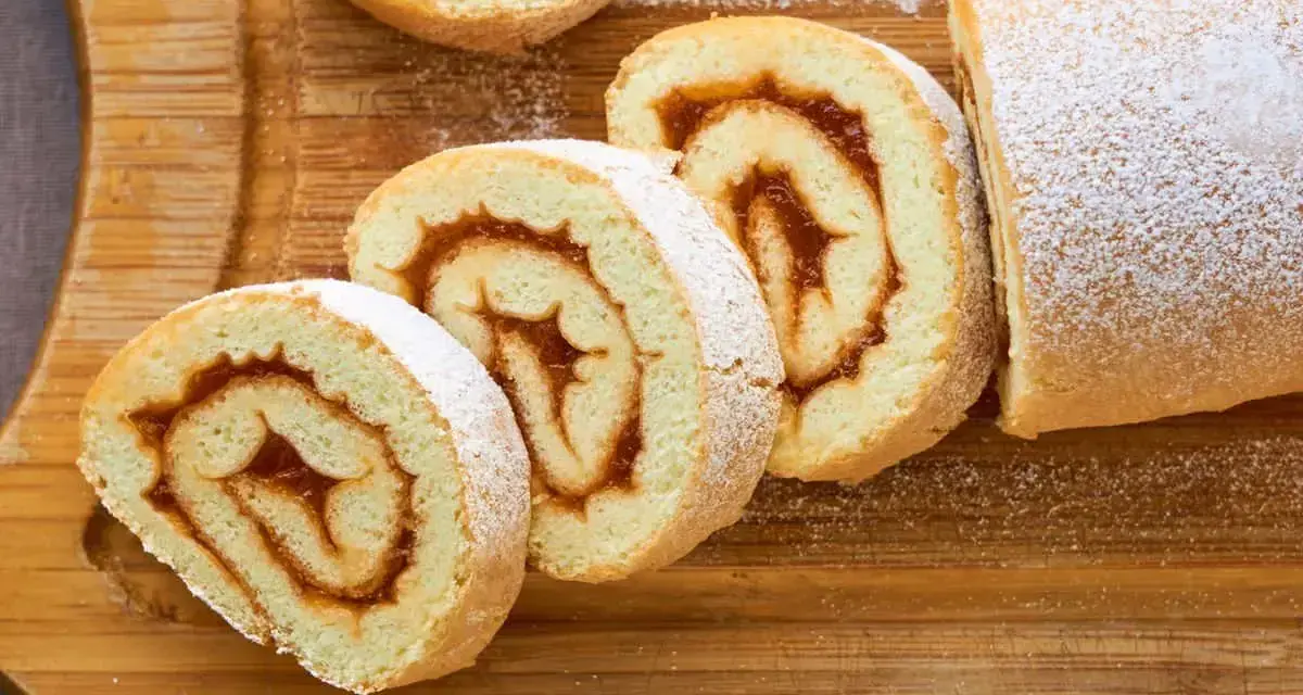 Rotoli di pasta biscotto 3 uova con ripieno di marmellata, spolverati di zucchero a velo su tagliere di legno.