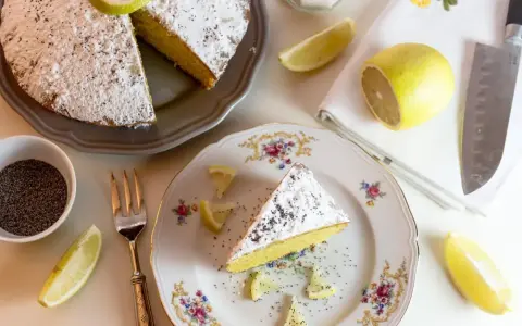Una fetta di babka cytrynowa, cosparsa di zucchero a velo e semi di papavero, con spicchi di limone e una ciotolina di semi.