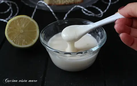 Una mano preleva con un cucchiaio una densa glassa al limone trasparente da una ciotola di vetro. Accanto, una fetta di limone.