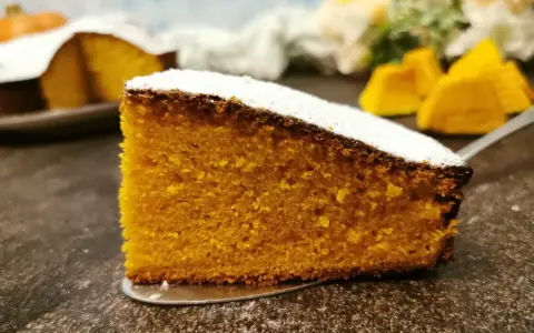 Una fetta di torta zucca senza glutine, soffice e dorata, cosparsa di zucchero a velo. Sullo sfondo, pezzi di zucca e un'altra torta.