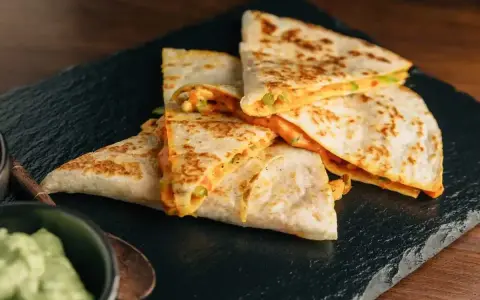 Deliziose quesadillas di pollo dorate e ripiene di formaggio filante, servite su una pietra scura con una ciotola di guacamole.