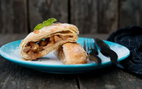Delizioso strudel pere cioccolato e amaretti, con ripieno di mele e un tocco di menta, su un piatto blu cosparso di zucchero a velo.