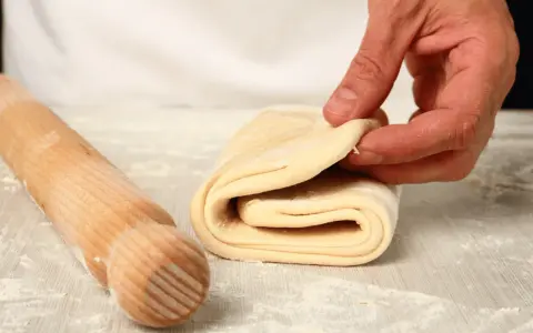 Mano che piega un impasto stratificato, pronto per diventare delizioso **ciasto francuskie**. Un mattarello è accanto.