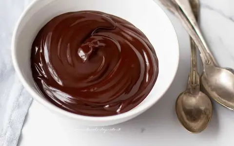 Una ciotola bianca piena di glassa al cioccolato e burro, lucida e invitante, con due cucchiai d'argento accanto.