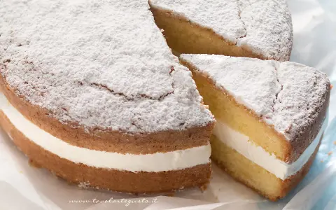 Una soffice torta paradiso con crema bianca, spolverata di zucchero a velo. Una fetta è stata tagliata, rivelando la deliziosa farcitura.