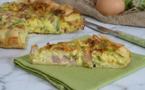 A slice of ham and zucchini quiche on a green napkin, with the rest of the quiche in the background.