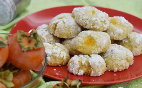 Biscotti ai cachi, dolcetti morbidi e profumati, cosparsi di zucchero a velo, accanto a cachi freschi.