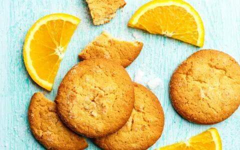 Biscotti all'arancia ricetta della nonna: dolci profumati e dorati, con spicchi d'arancia fresca su un tavolo di legno azzurro.