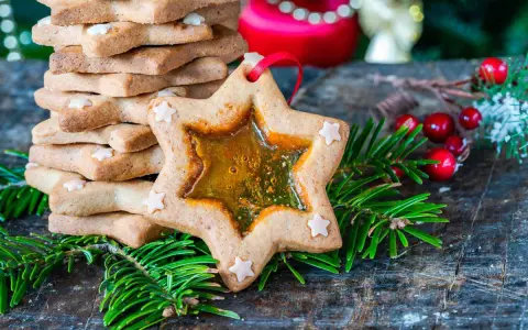 Biscotti a forma di stella con centro trasparente, decorati con piccoli fiocchi di neve, circondati da rami di abete e bacche rosse.
