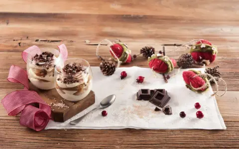 Due bicchieri di tiramisù Galbani, decorati con cioccolato e nastri rossi, su un tavolo di legno con decorazioni natalizie.