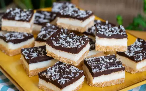 Deliziosi quadrotti cioccolato e cocco, stratificati con base croccante, crema bianca e copertura fondente, spolverati con cocco grattugiato.