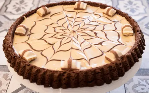 Torta con base croccante al cacao, ripiena di crema chiara e decorata con cioccolato fondente e pezzi di Kinder Bueno White.