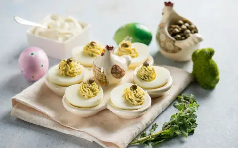 Uova ripiene vegetariane con ceci, guarnite con capperi e erbe aromatiche, su un piatto decorato con galline e uova pasquali.