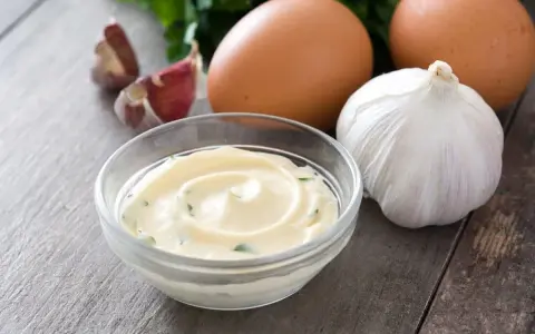 Una ciotola di cremoso sos aioli, con uova, aglio e prezzemolo sullo sfondo.