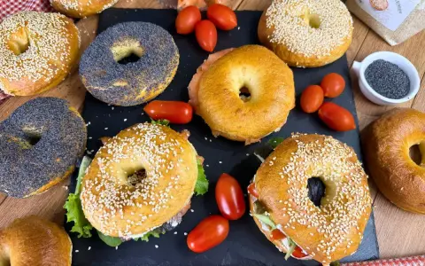 Varietà di bagel dolci e salati, alcuni con semi di sesamo, altri con semi di papavero, accompagnati da pomodorini e salmone affumicato.
